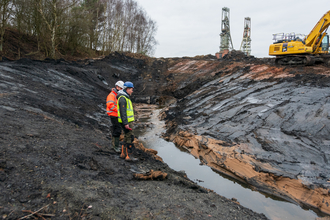 Colliery pitheads with a big digger clearing a river bed with people onlooking